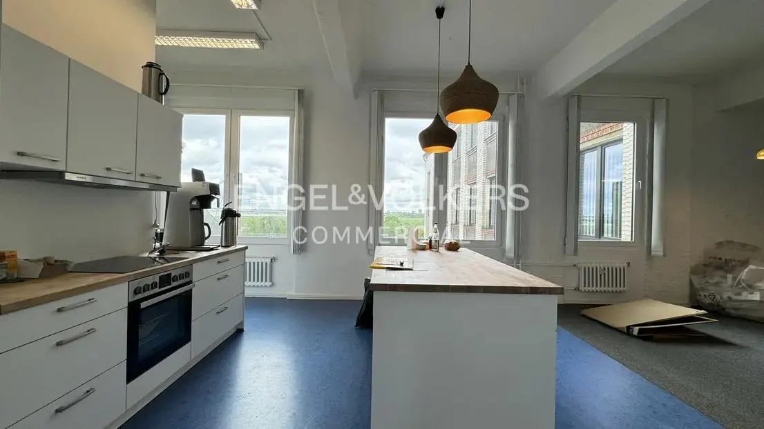 Bright office kitchen with white cabinets, wood countertops, and a central island under two pendant lights. Large windows offer natural light.