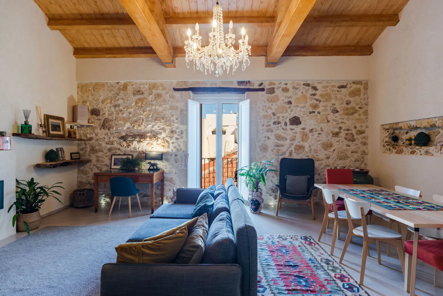 Living room with stone wall, wood ceiling, and chandelier. Gray sofa, desk, dining table, and patterned rug. Balcony view.