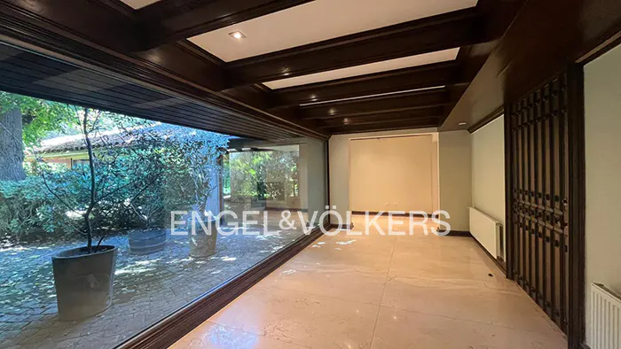 Interior view of a room with marble floors, dark wood trim, and a large window overlooking a garden. The Engel & Völkers logo is visible on the window.