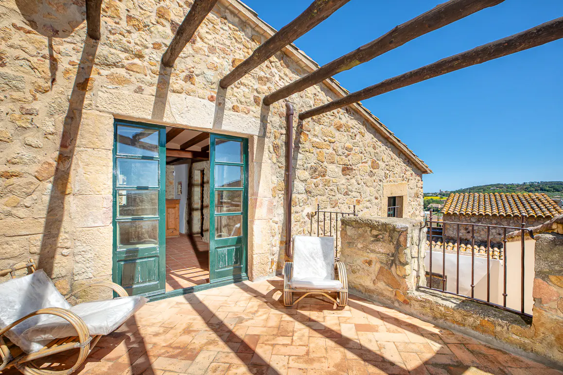 Stone house terrace with open green doors, wicker chairs, and a wooden pergola against a clear blue sky.