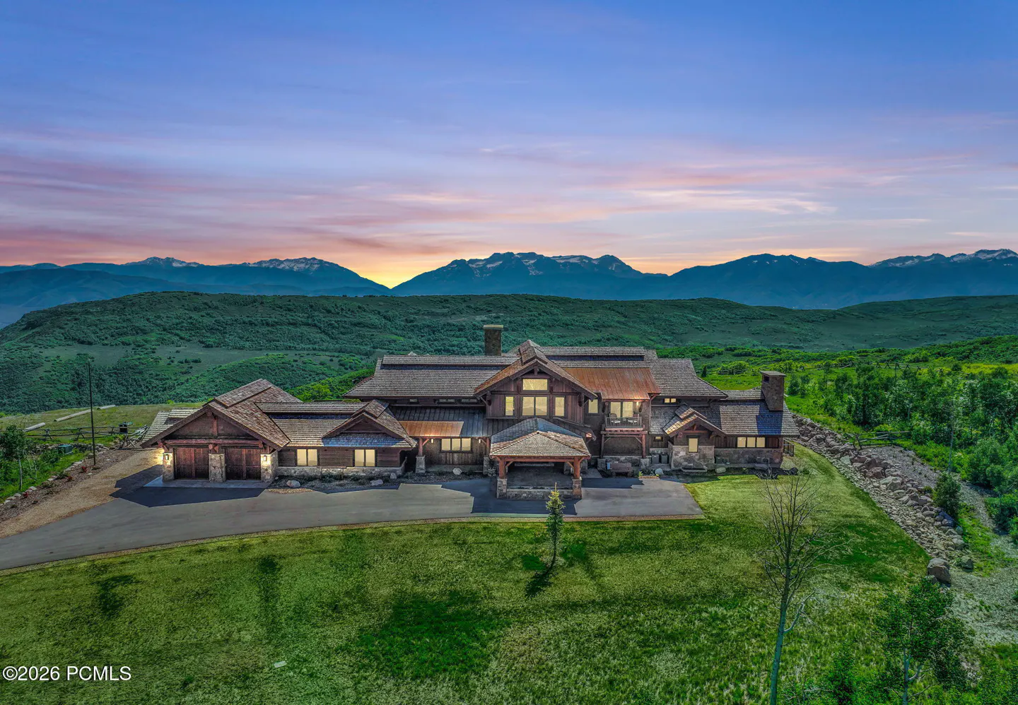 A large, brown, luxury home sits on a green lawn with mountains in the background at sunset.