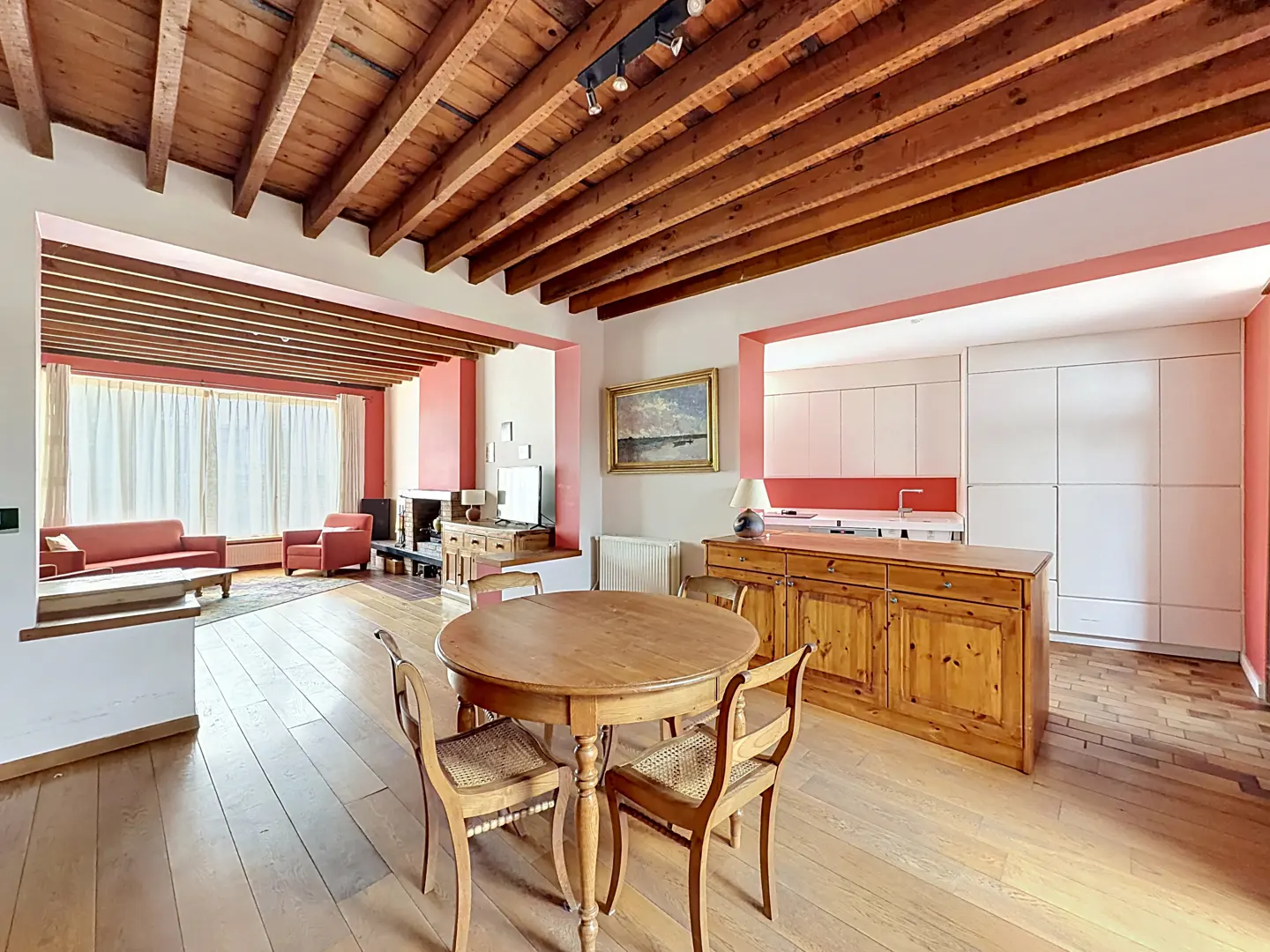 Open-plan living area with wooden ceiling beams, a round dining table, kitchen island, and a lounge with red furniture.