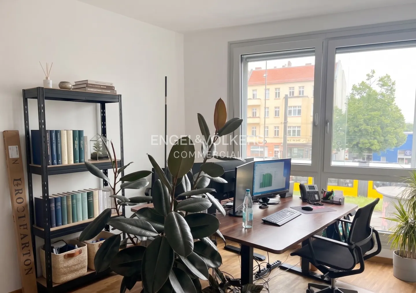 Office space with a desk, computer, chair, and bookshelf. A large window overlooks a city street.