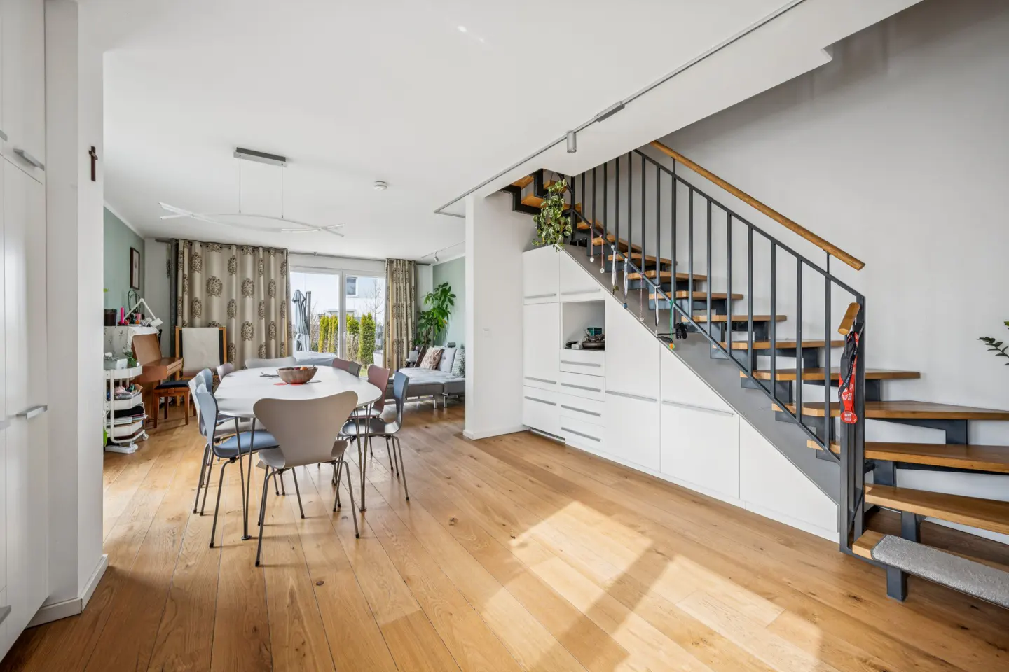 Open-plan living area with light wood floors, a dining table, and a modern staircase with white built-in storage below.