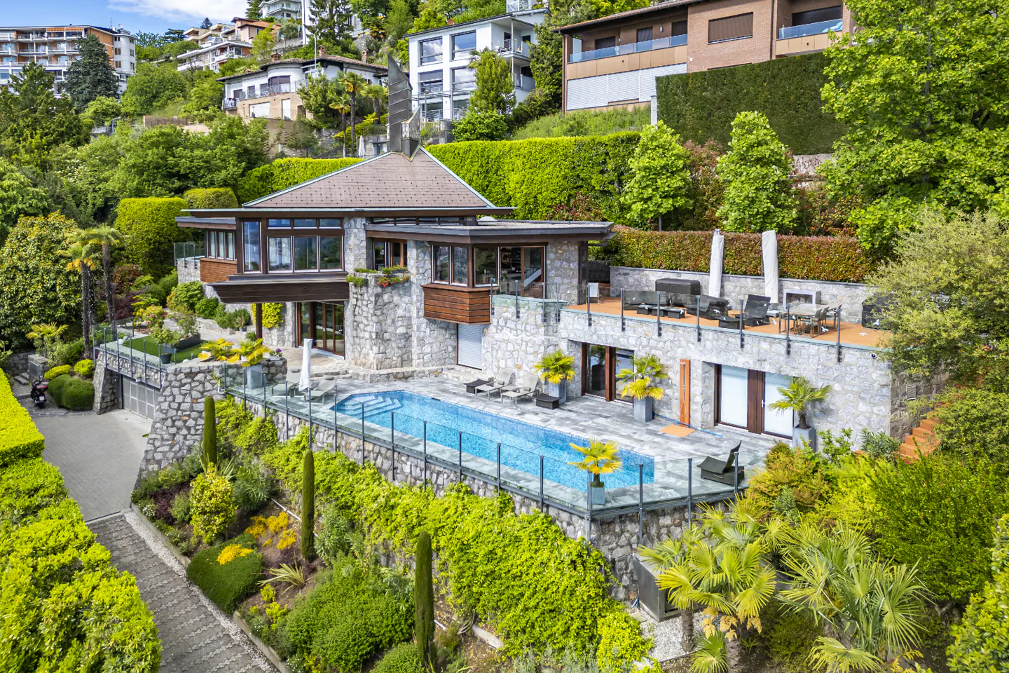 Luxury stone house with a pool on a green hillside. The pool is blue and surrounded by a glass fence. A patio has chairs and a grill.