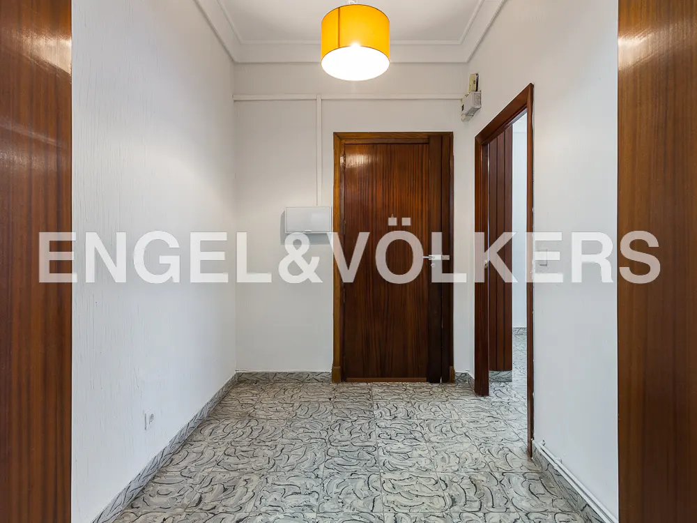 A bright hallway with white walls, patterned tile floor, and two dark wood doors. A yellow lampshade hangs from the ceiling.