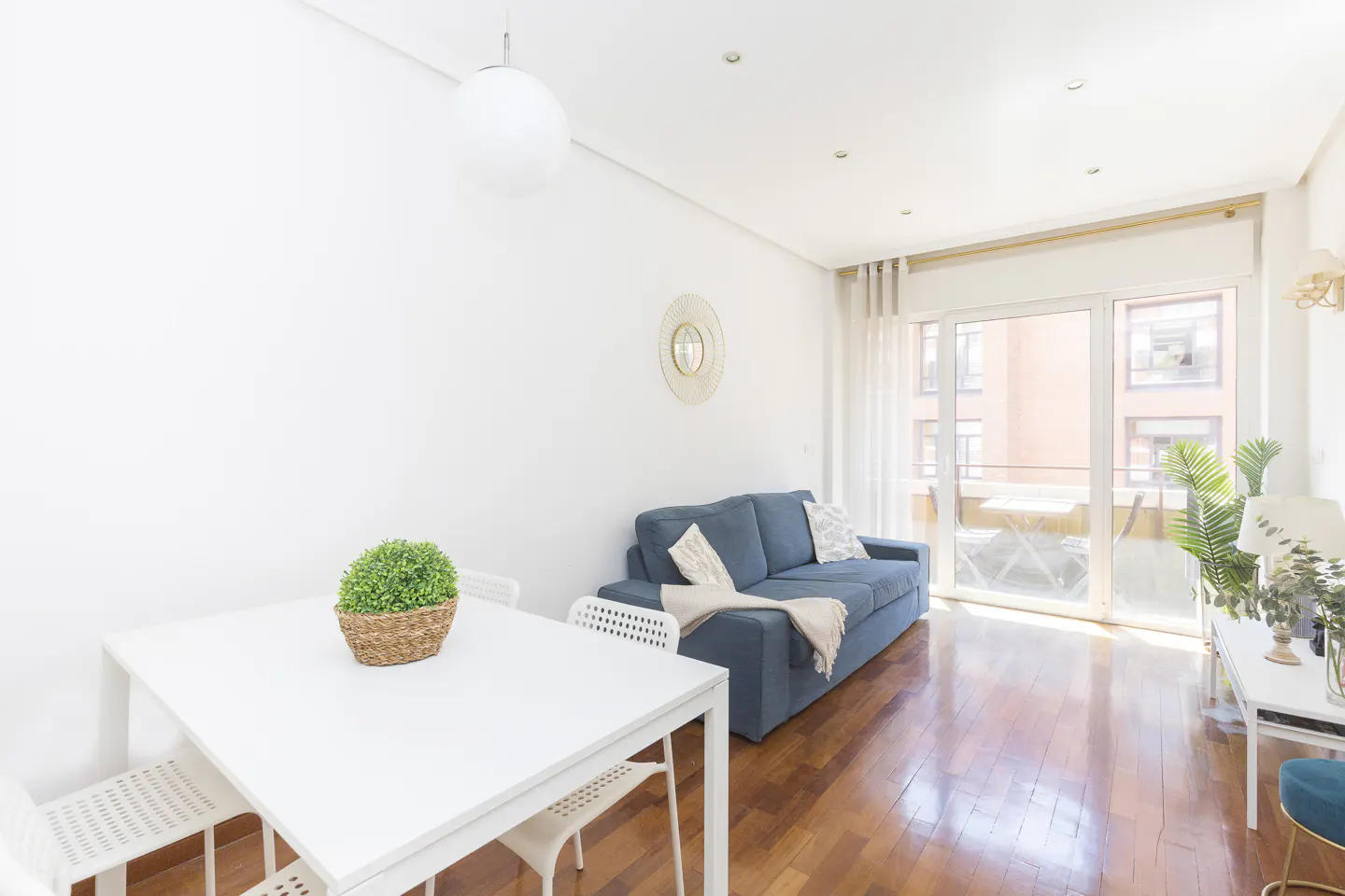 Bright living room with white walls, wood floors, and a blue sofa. A white table with chairs sits in the foreground. Balcony doors let in natural light.