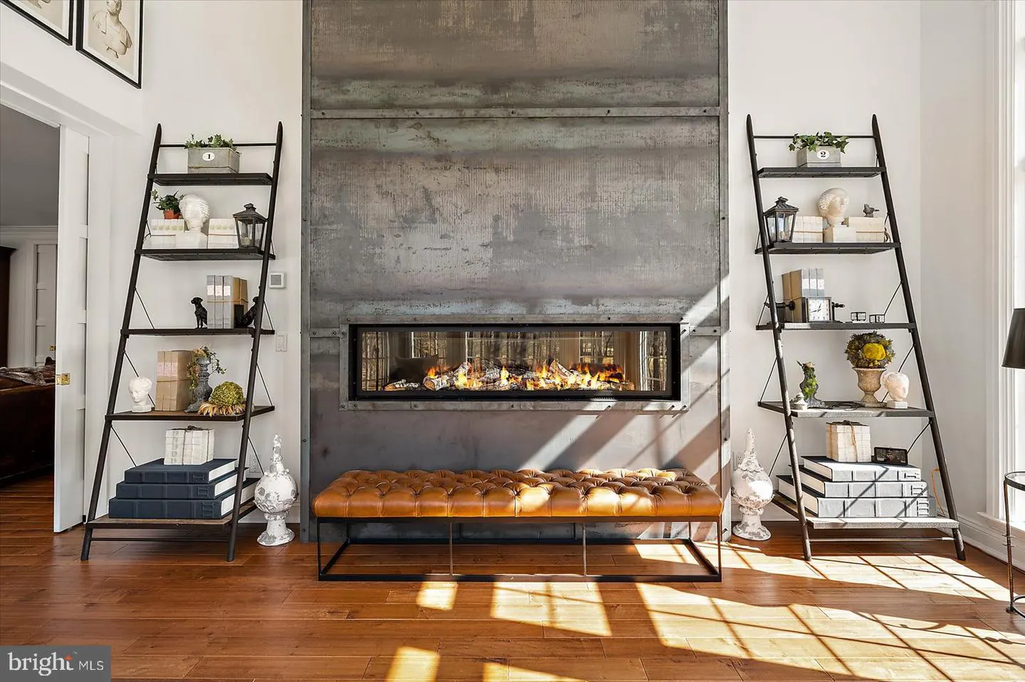 Living room with a long, modern fireplace, flanked by ladder shelves. A tufted leather bench sits in front of the fireplace. Hardwood floors.