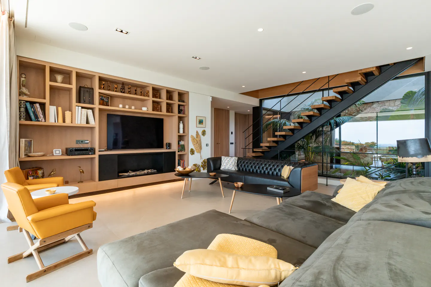A modern living room with a gray sectional sofa, yellow armchairs, and a black leather sofa. A staircase is visible through a large window.