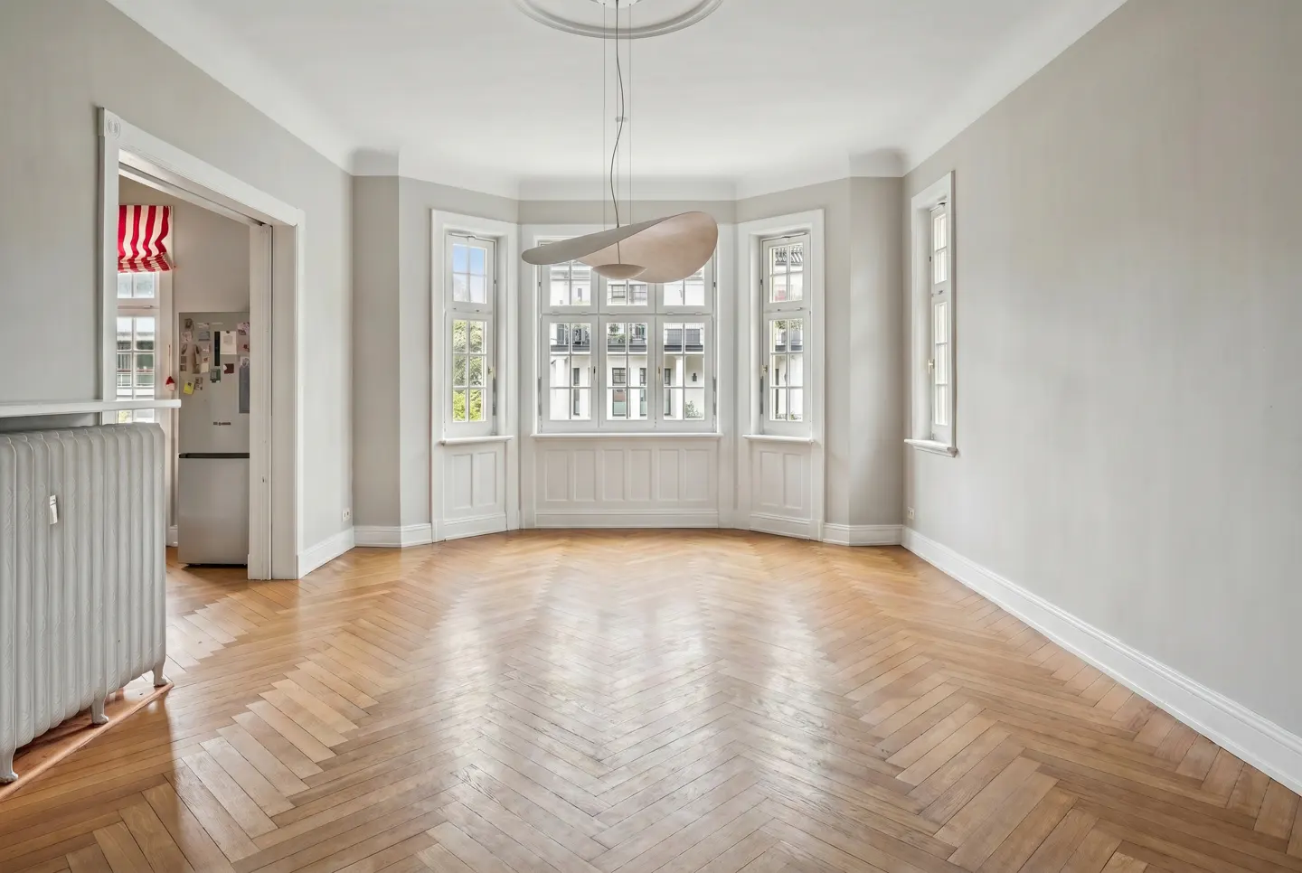 Bright, empty room with herringbone wood floors, gray walls, and white trim. A modern light fixture hangs from the ceiling. Windows offer a view of buildings.