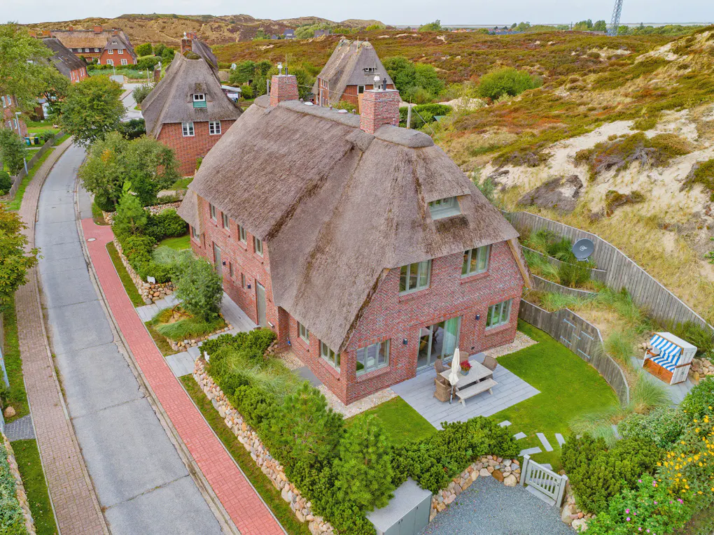 Aerial view of a red brick house with a thatched roof, green trim, and a well-manicured lawn. A road and other houses are visible.