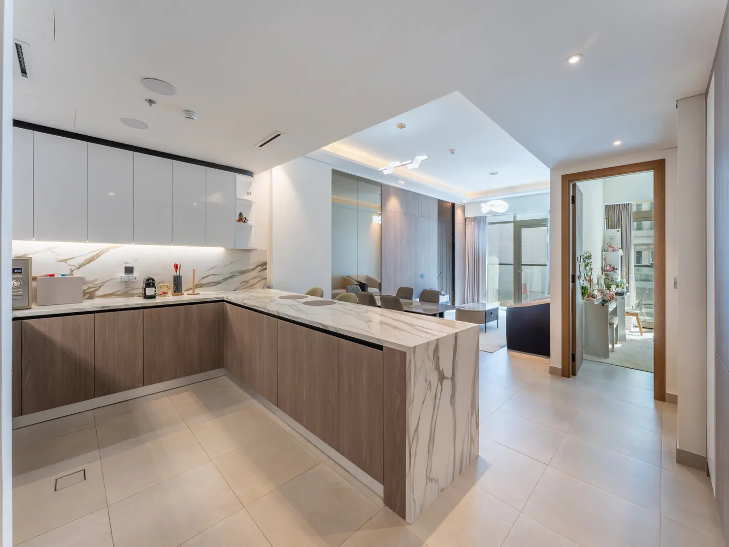 A bright, modern kitchen with white cabinets, marble countertops, and wood-paneled lower cabinets, open to a living area with a dining table.
