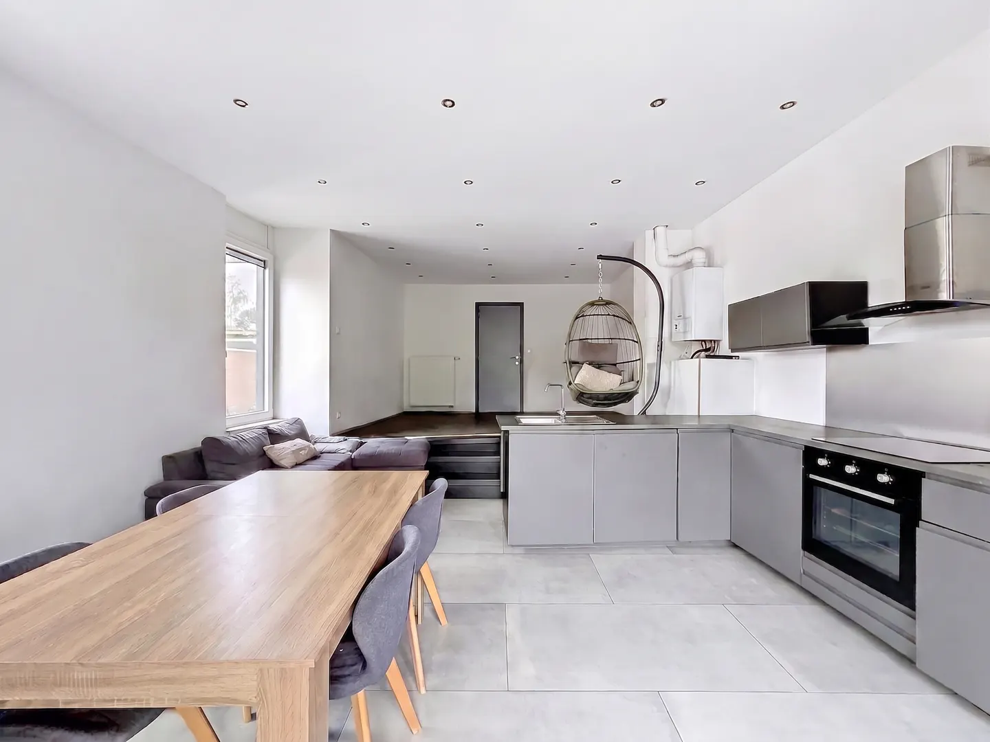 Open-concept living space with a gray kitchen, wood table, gray sofa, and hanging chair.
