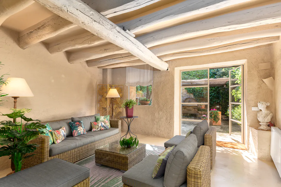 Living room with exposed beams, wicker furniture, and a view to a lush garden through an open glass door.
