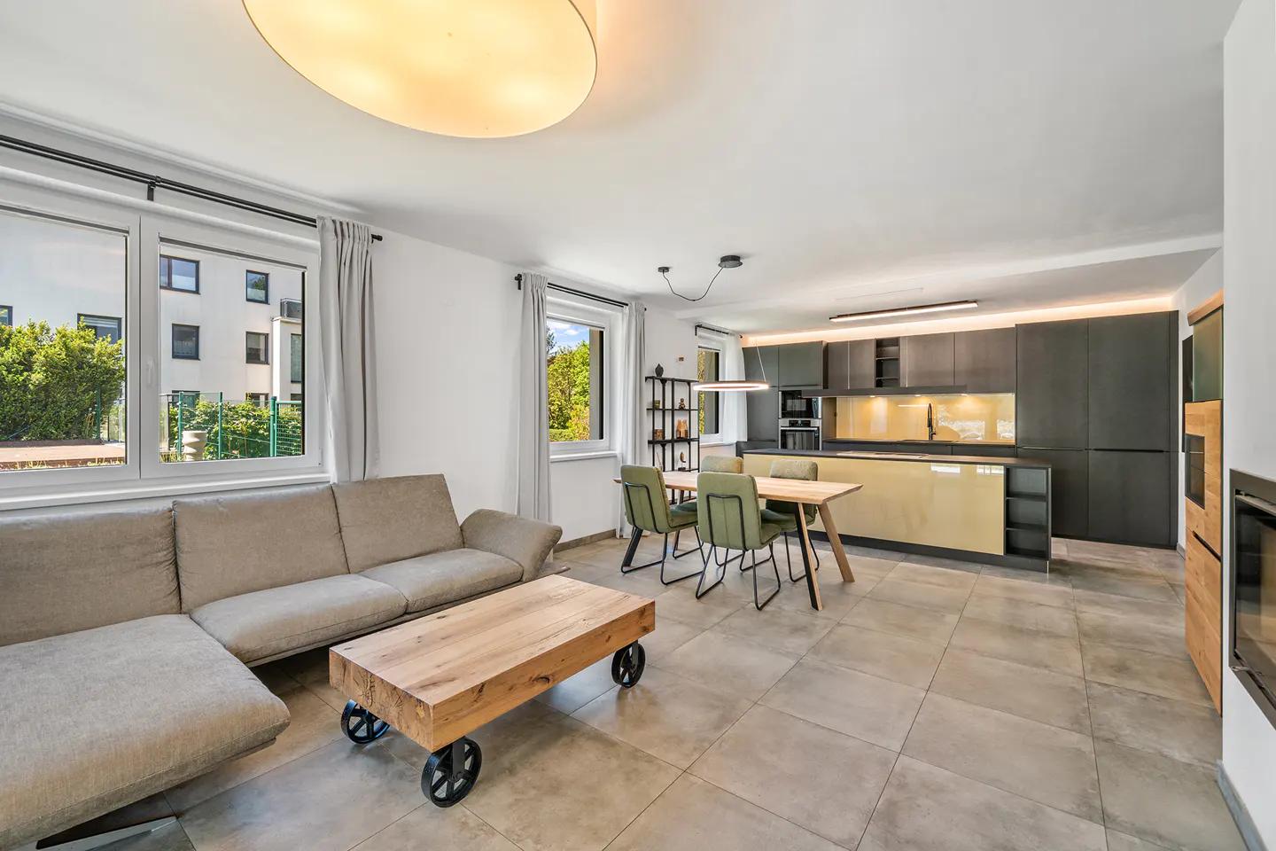 Open-concept living space with a beige sofa, wood coffee table on wheels, dining table with green chairs, and a modern black and gold kitchen.