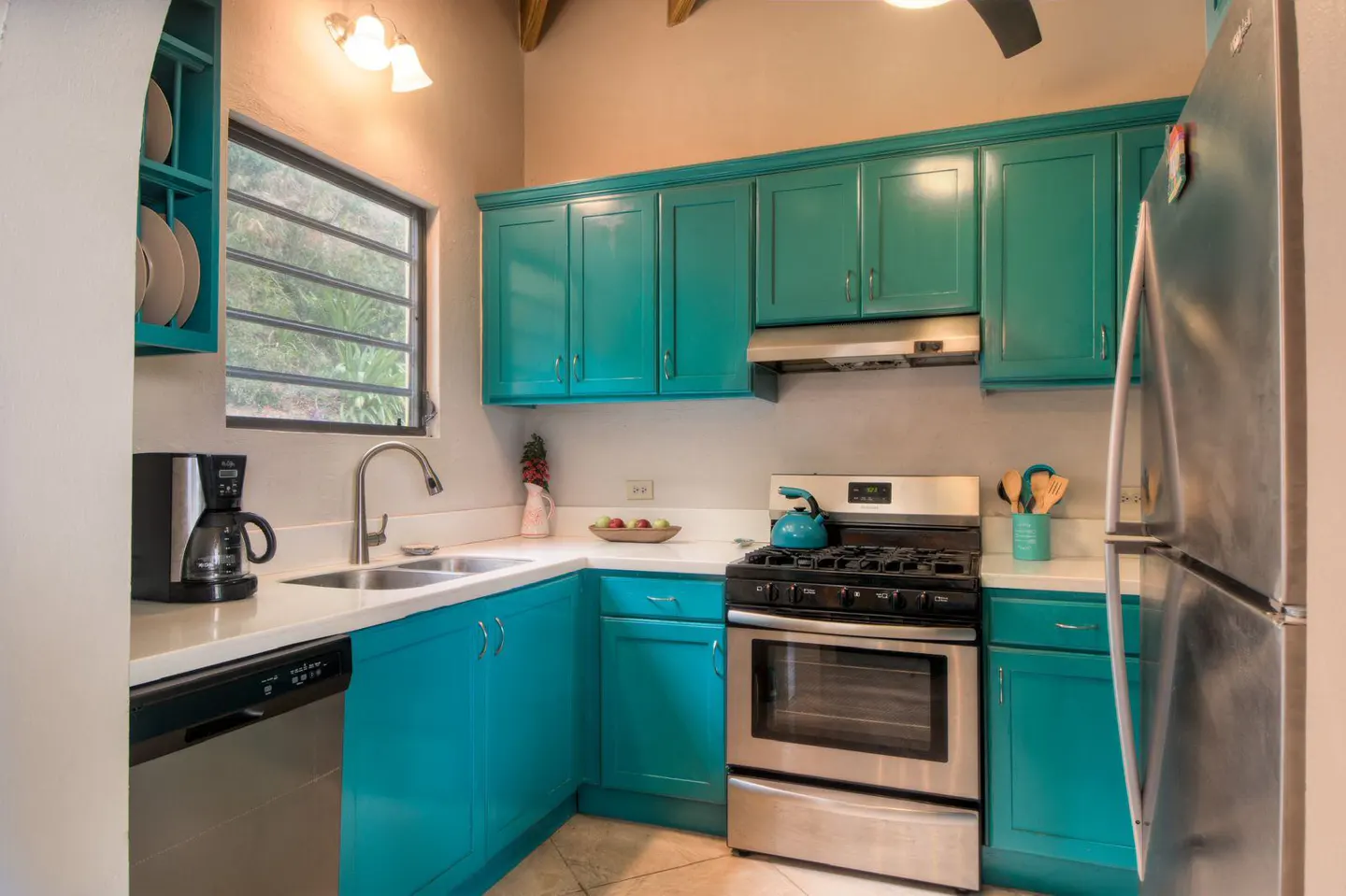 A kitchen with turquoise cabinets, stainless steel appliances, and white countertops. A window is above the sink.