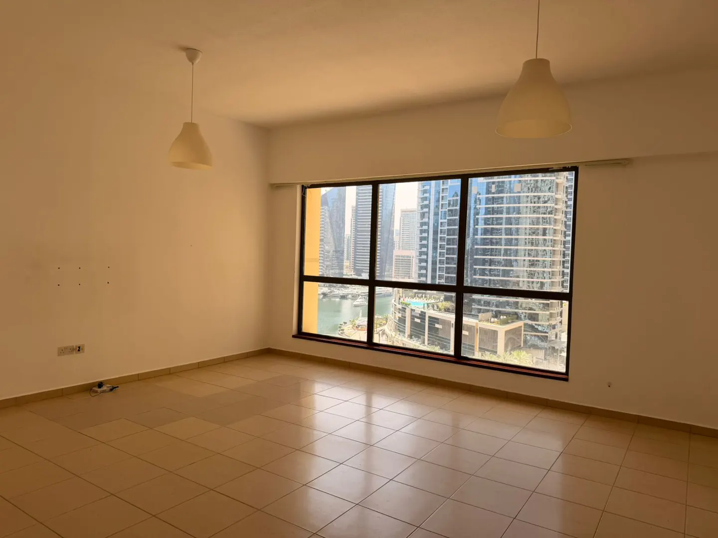 Empty room with beige tile floor, white walls, and a large window showing a city skyline. Two pendant lights hang from the ceiling.