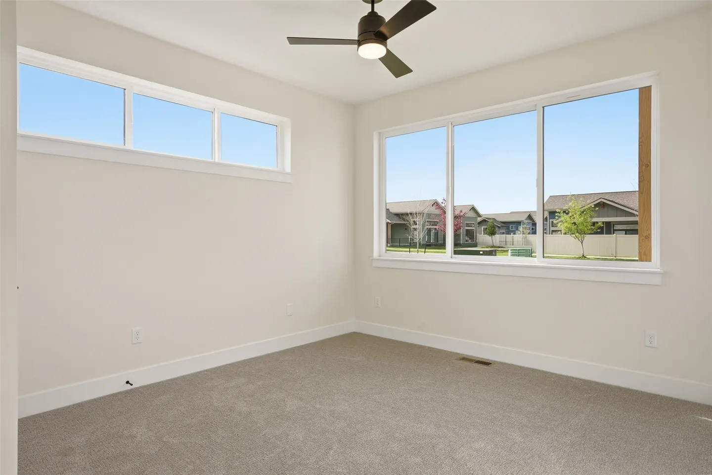 Bright, empty room with gray carpet, white walls, and a dark ceiling fan. Large windows offer a view of houses and a blue sky.