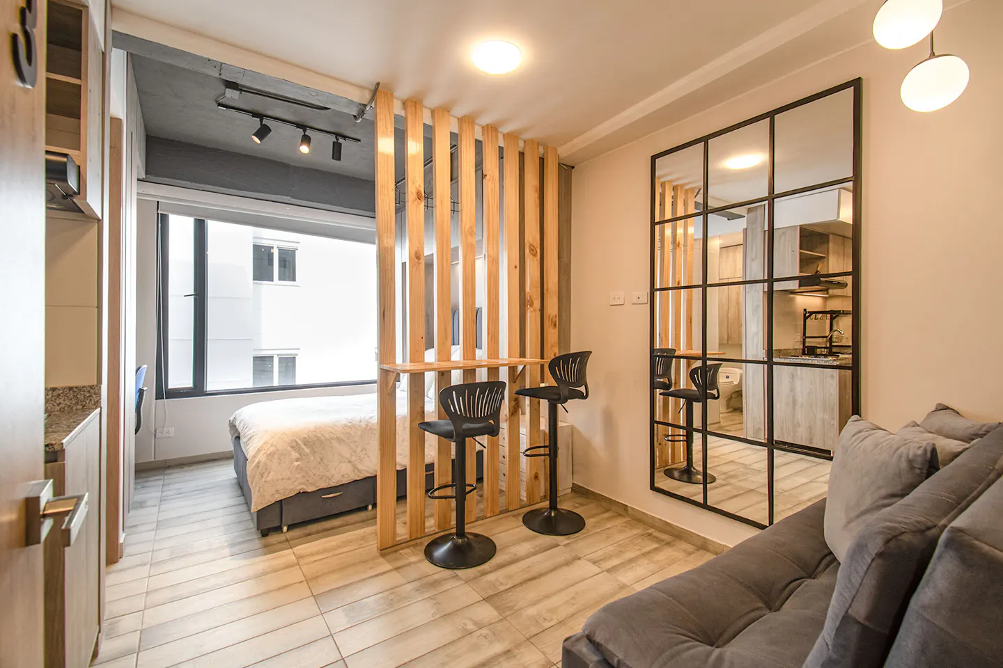Studio apartment with a gray sofa, two black bar stools at a wooden divider, and a large window with a bed visible.