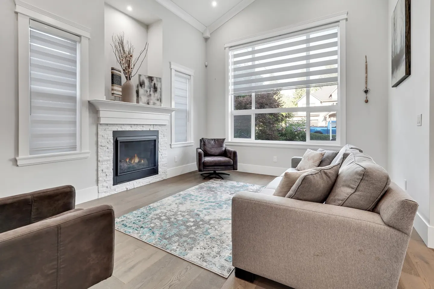 Bright living room with a stone fireplace, a gray sofa, and a patterned rug on a wood floor. Large windows with blinds let in natural light.