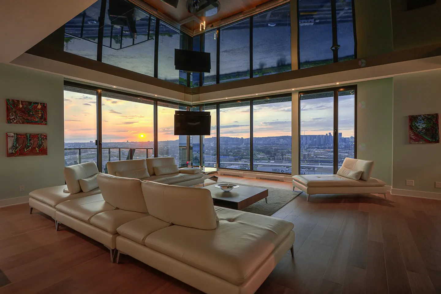 Living room with a large white sofa, hardwood floors, and floor-to-ceiling windows showing a sunset over a city.