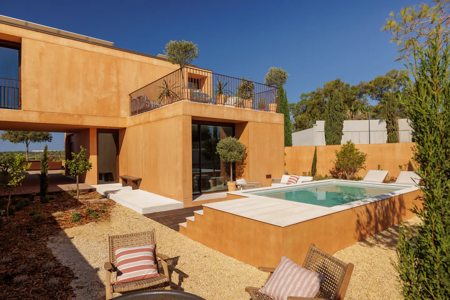 Modern orange stucco home with a pool and gravel yard. Lounge chairs and trees surround the pool. Balcony with plants on the roof.