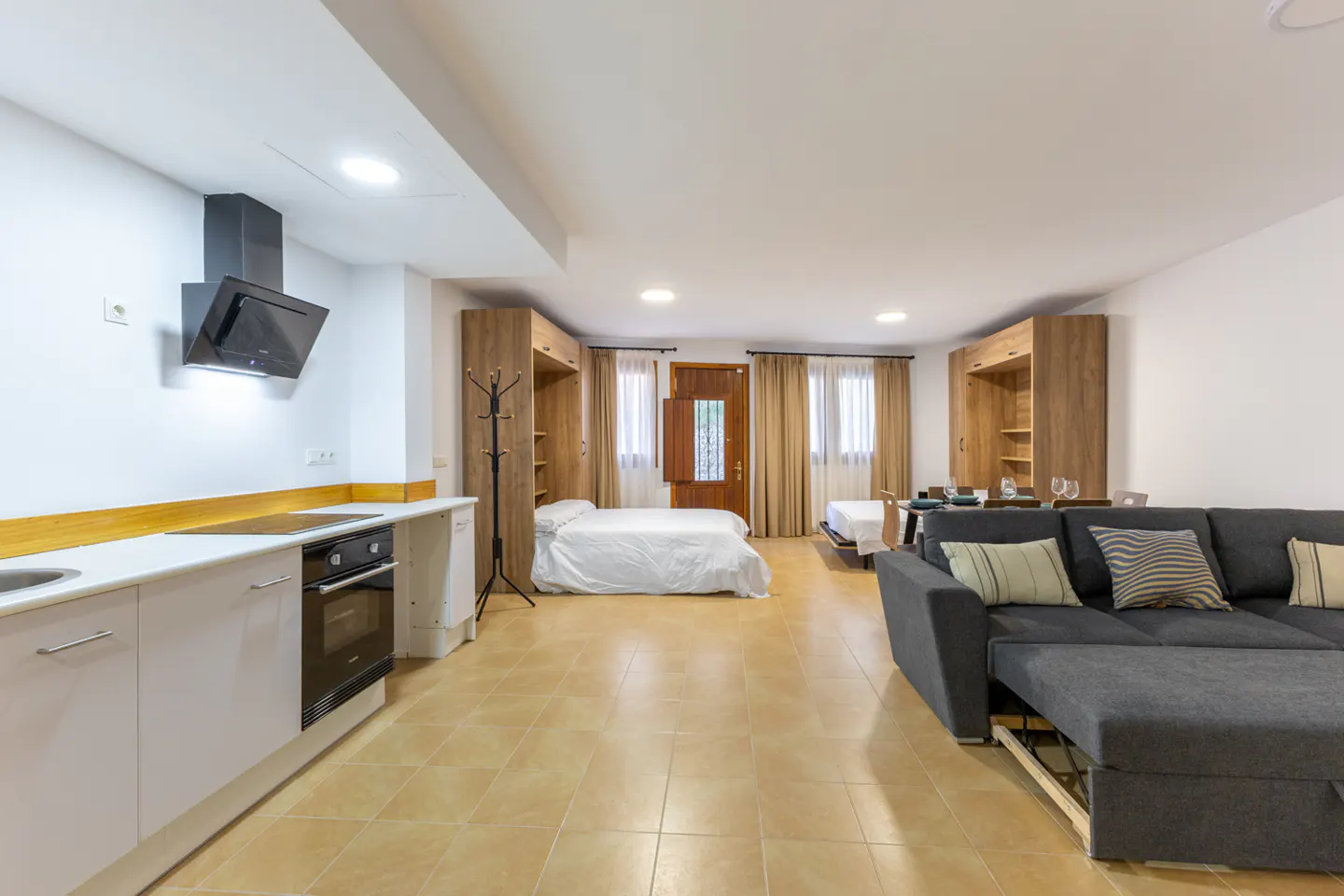 Studio apartment with a kitchen, two beds in wooden cabinets, and a gray sofa with pillows. The floor is tiled.