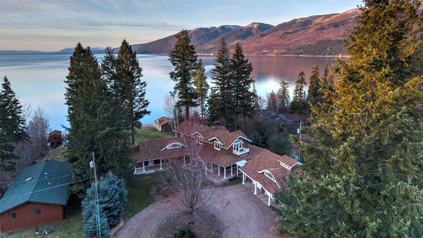 Aerial view of a large brown house with a lake and mountains in the background.