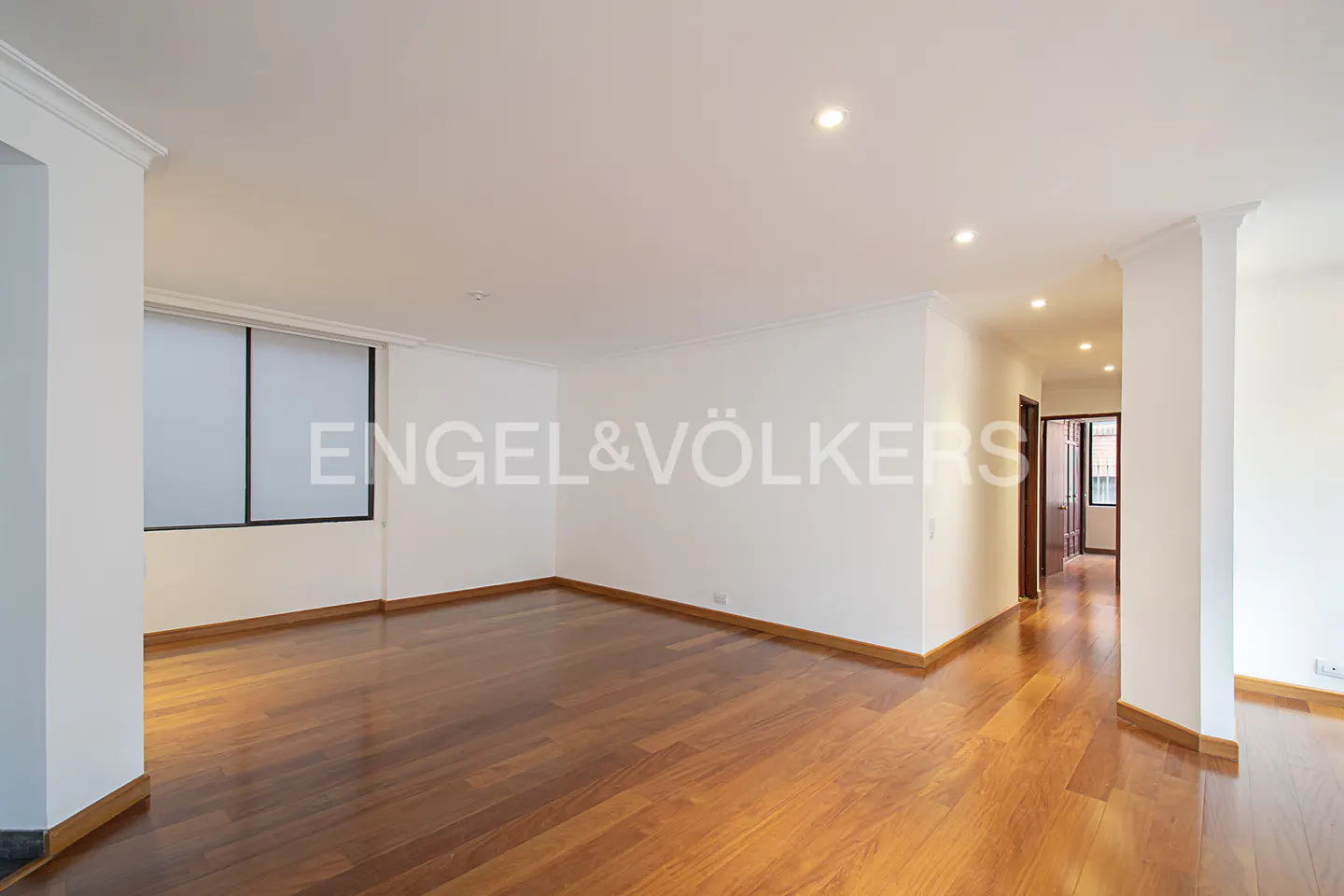Empty living room with hardwood floors, white walls, and a window. A hallway leads to other rooms. Engel & Völkers logo is visible.