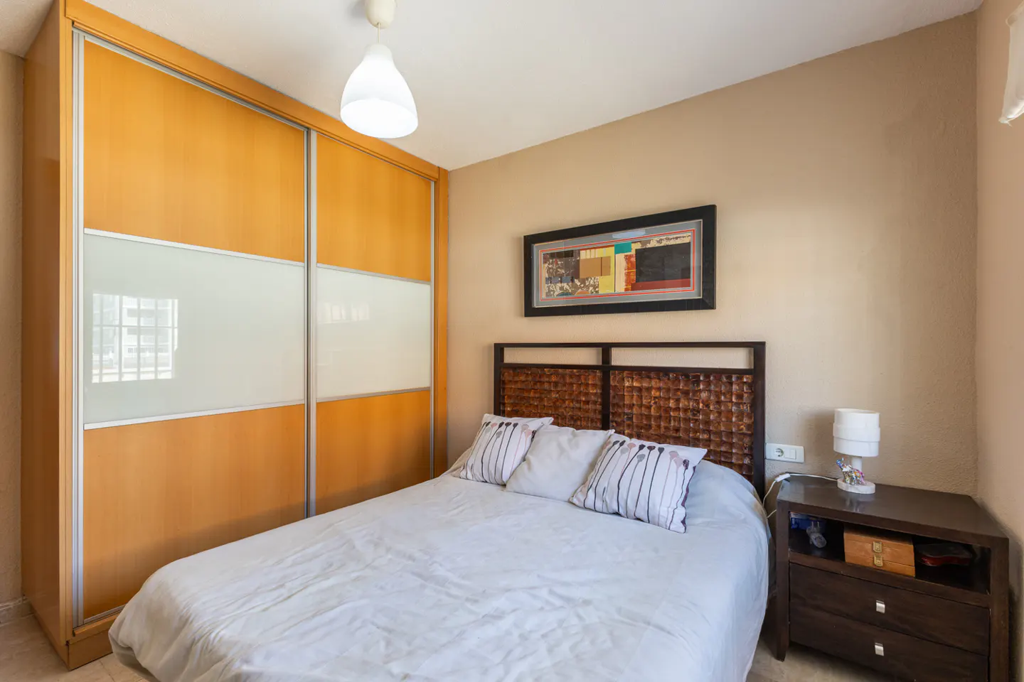 Bedroom with a double bed, woven headboard, bedside table, and a large sliding door wardrobe. The walls are beige.