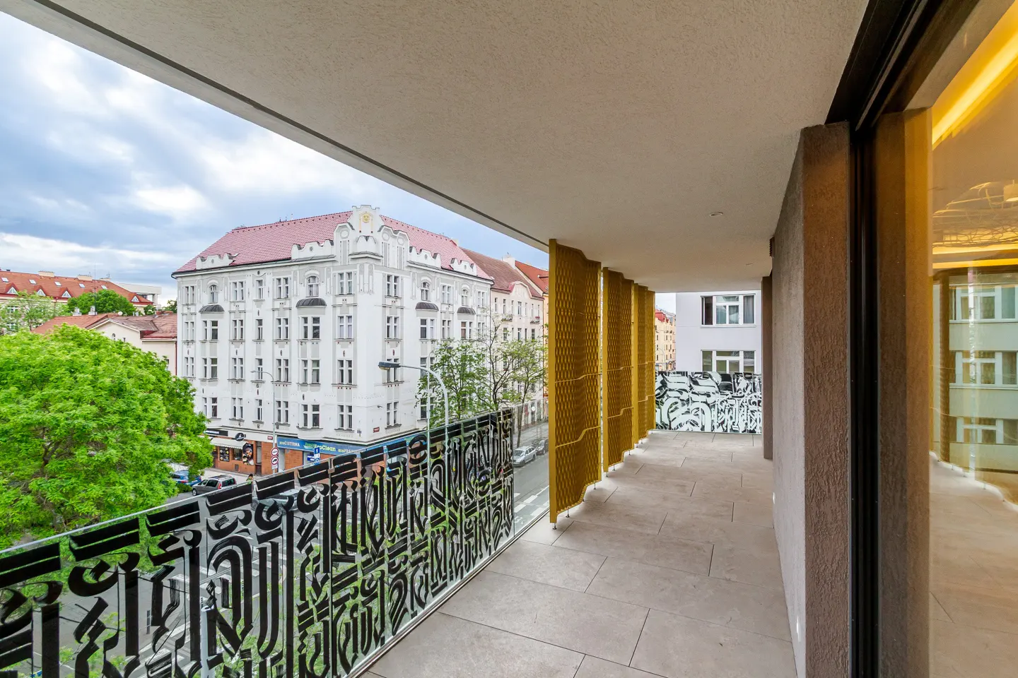 View from a balcony with black decorative railings, gold mesh screens, and stone flooring, overlooking a city street with buildings and trees.