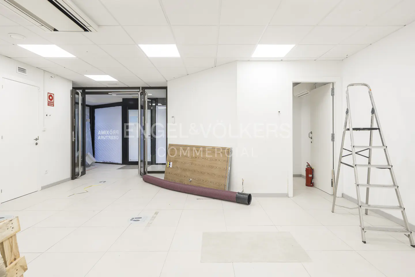 Empty white commercial space under renovation with a metal stepladder, a wooden panel, and a red fire extinguisher.