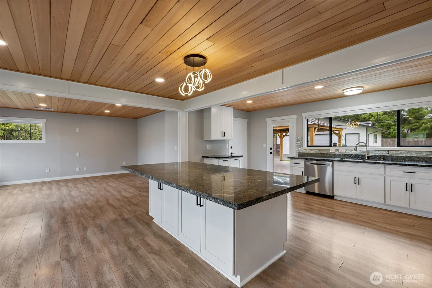 A bright, open kitchen with a wood plank ceiling, white cabinets, and a large island with a granite countertop. A window overlooks the backyard.