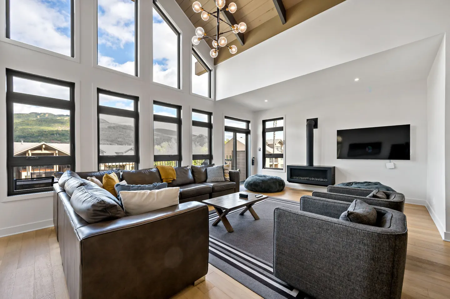 Bright living room with a brown leather sofa, gray chairs, and a modern fireplace. Large windows offer mountain views.