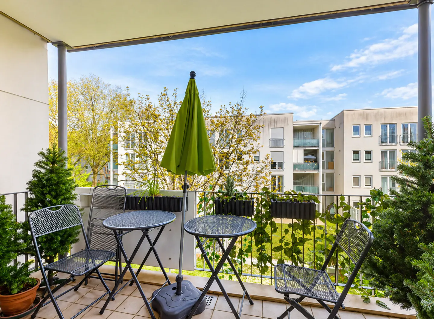 Balcony view with metal chairs, tables, and a green umbrella. Buildings and blue sky are visible in the background.