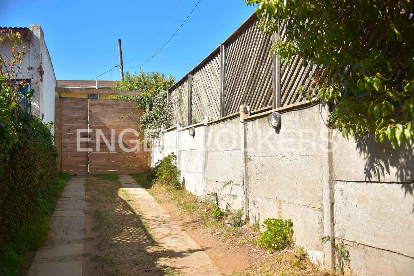 Exterior view of a driveway leading to a wooden gate, bordered by a concrete wall with a lattice fence.