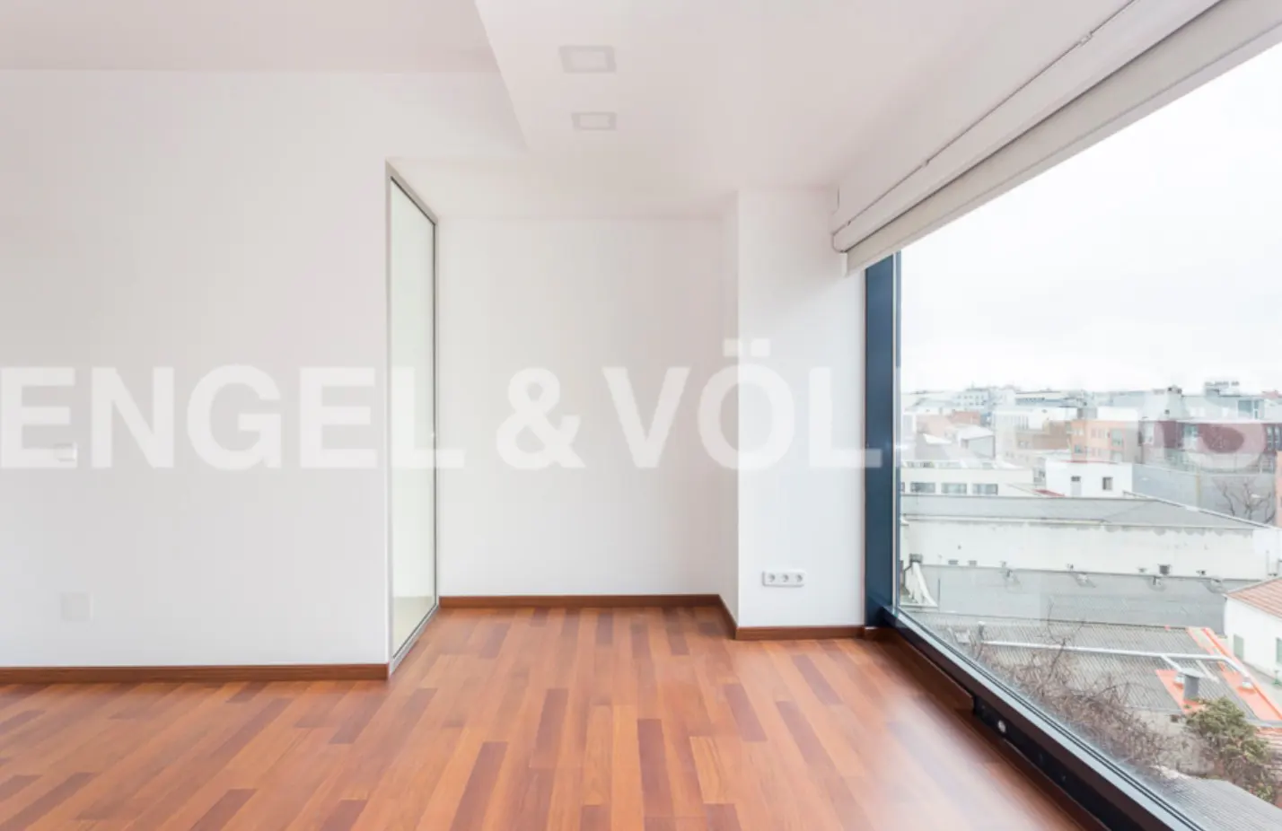 Bright, empty room with wood floors, white walls, and a large window overlooking a city. Engel & Völkers logo on the wall.