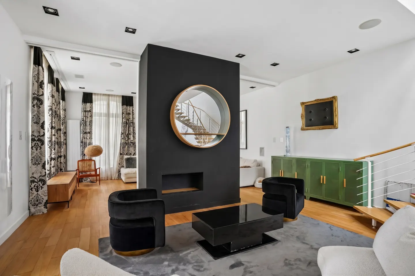 Living room with wood floors, black accent wall with round mirror, black chairs, and green cabinet.