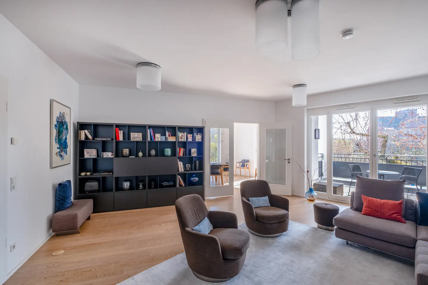 Bright living room with hardwood floors, a gray sofa, brown swivel chairs, and a large black bookcase. Balcony with outdoor furniture visible.