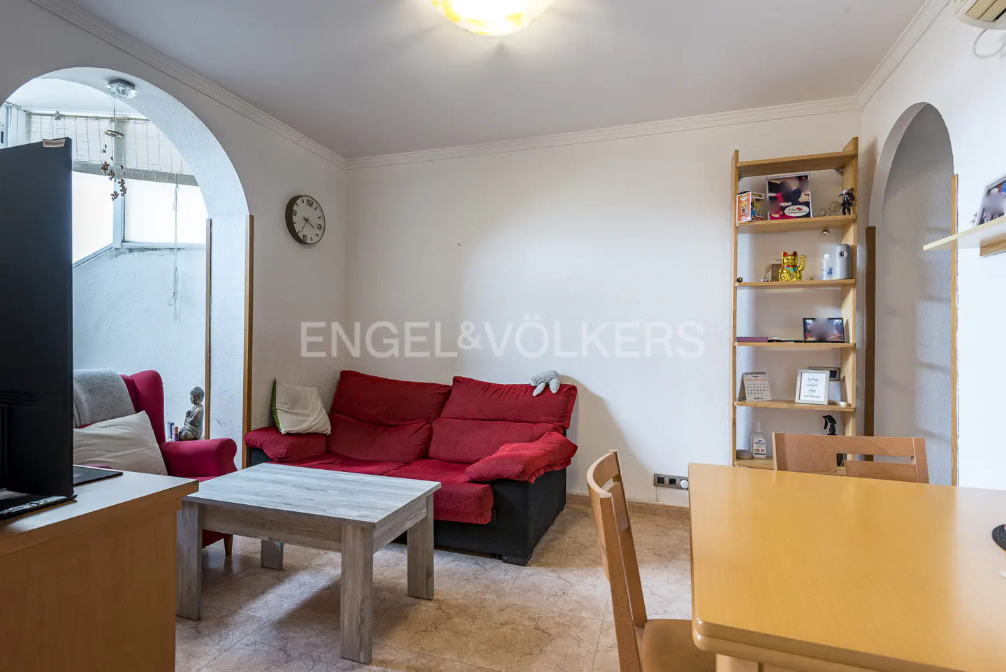 Living room with a red sofa, wooden coffee table, dining set, and a tall shelving unit against a white wall.