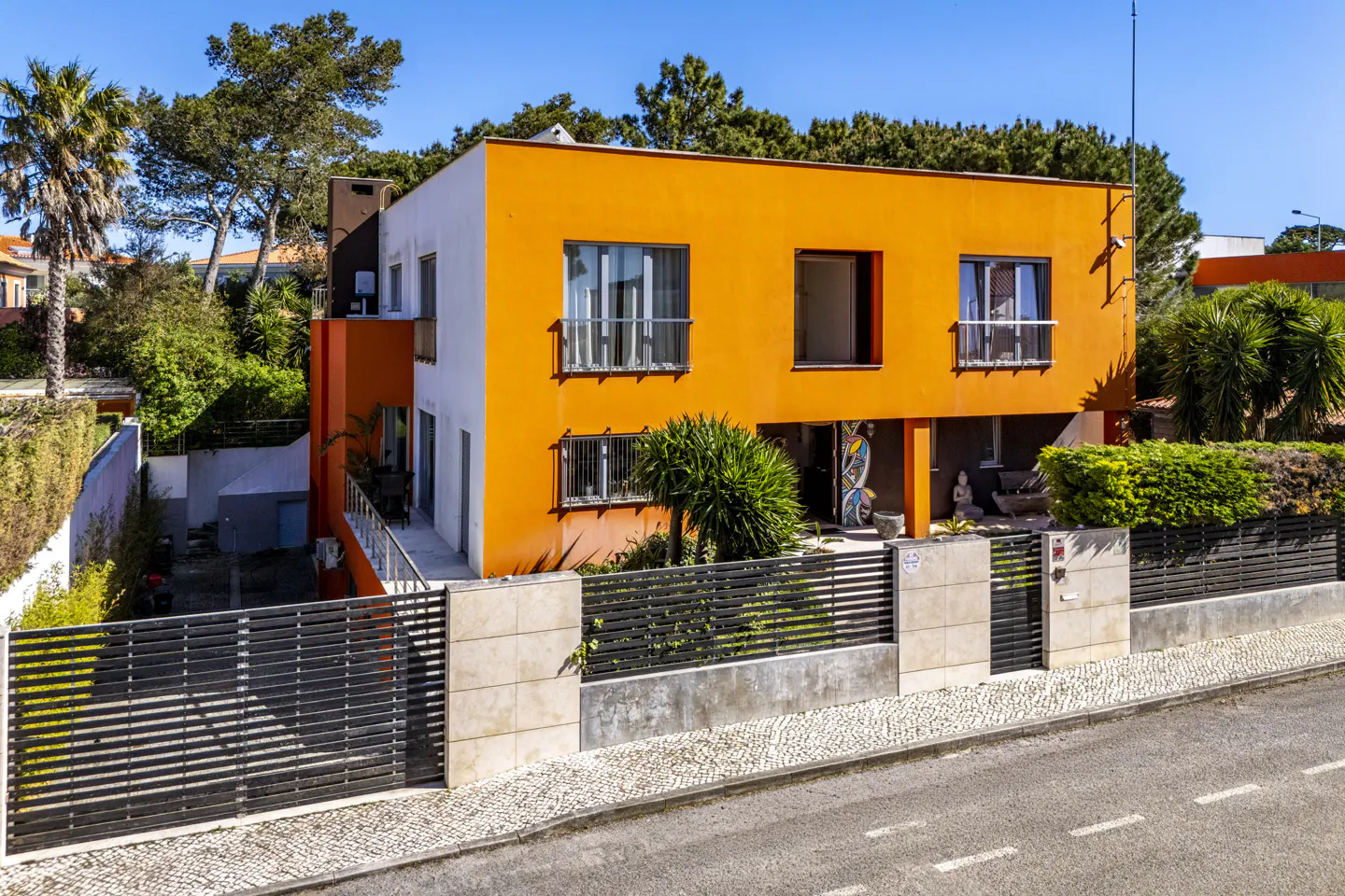 Two-story modern house with orange and white exterior, black fence, and green trees under a clear blue sky.