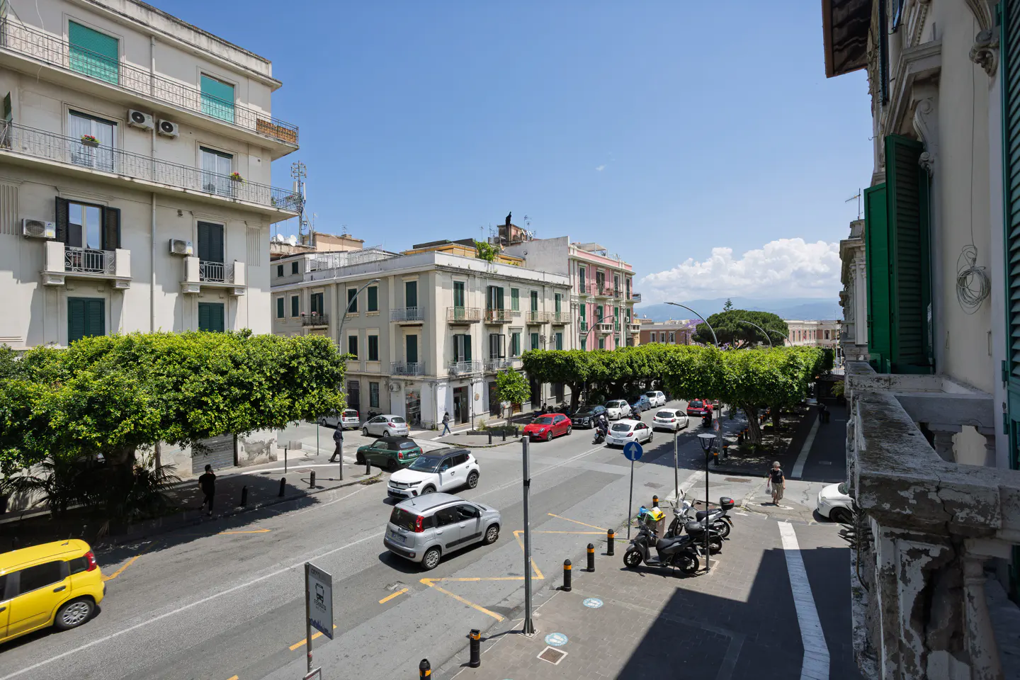 View from a balcony overlooking a sunny Italian street with historic buildings, lush green trees, and light traffic.