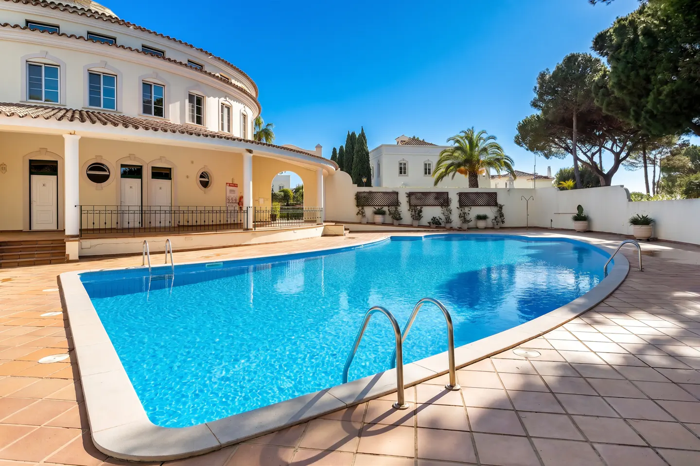 Outdoor pool with blue water and metal ladder, surrounded by a tiled patio and a cream-colored building with arched openings.