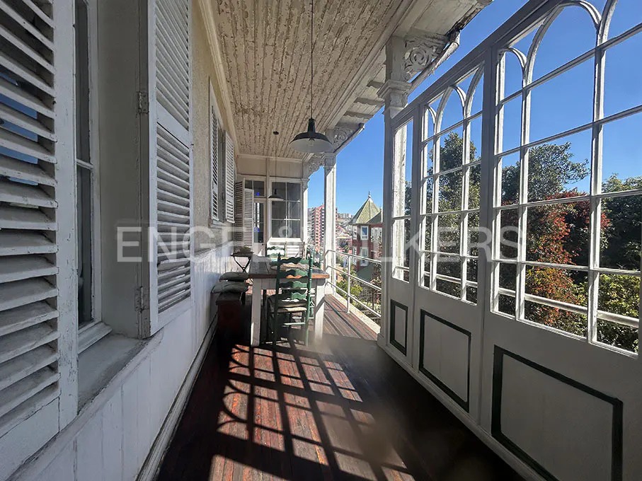 A long, narrow balcony with white walls, shutters, and arched windows. A table and chairs sit on the wooden floor, with a view of trees and buildings.
