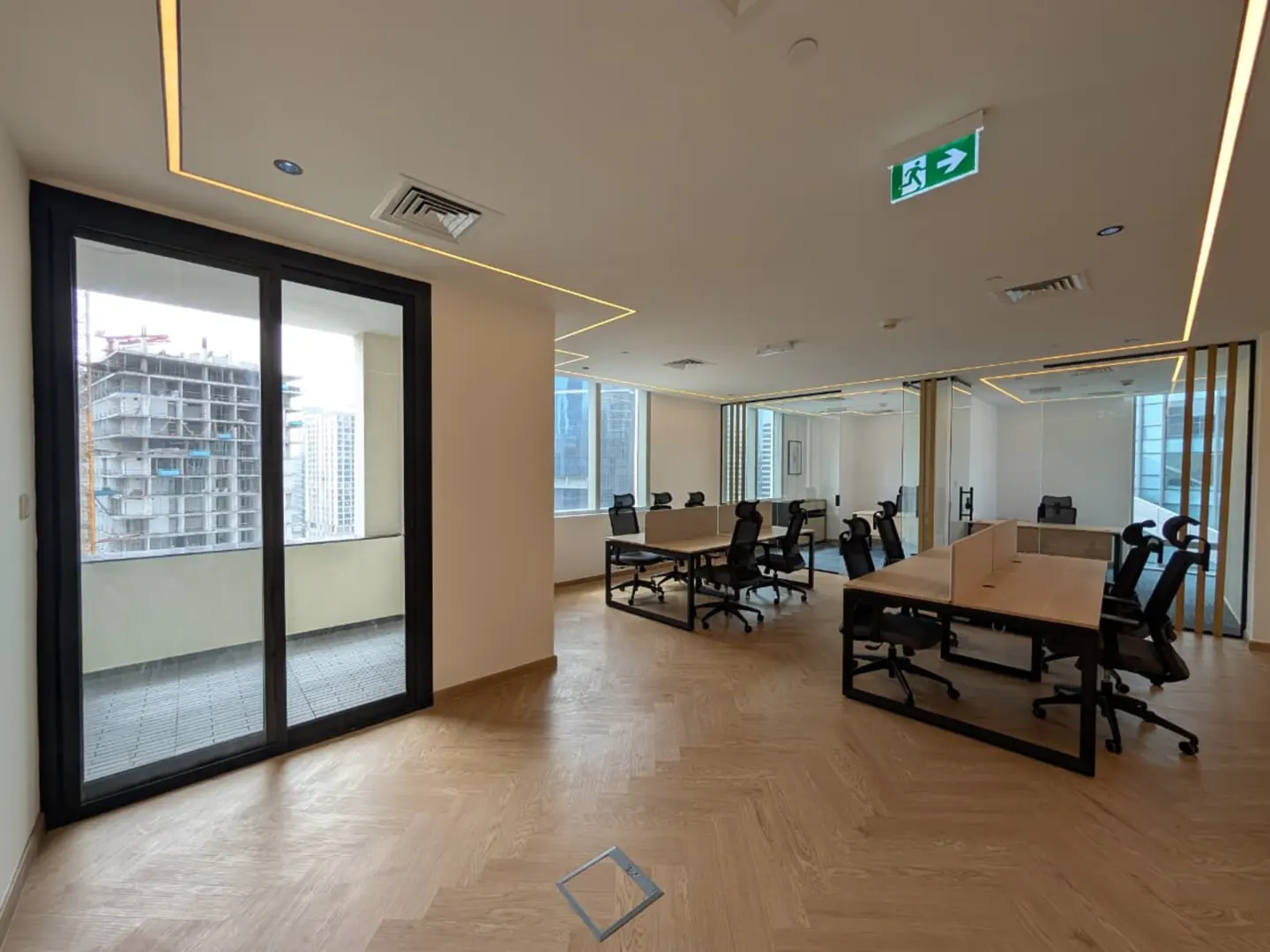 Bright, modern office space with desks, black chairs, and a large window overlooking a city construction site. Light wood floors and an exit sign are visible.