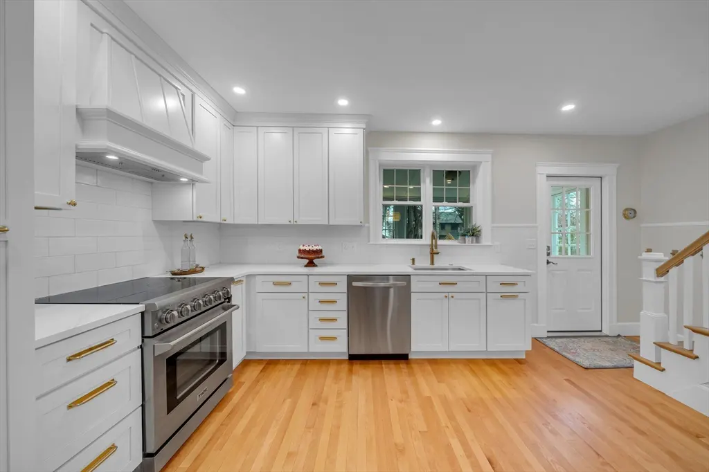 Bright, modern kitchen with white cabinets, gold hardware, stainless steel appliances, and hardwood floors. A white door and staircase are visible.