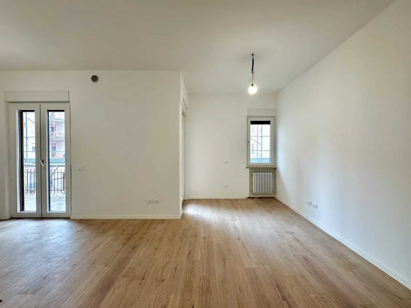 Bright, empty room with light wood floors, white walls, and a window. A glass door leads to a balcony. A single lightbulb hangs from the ceiling.