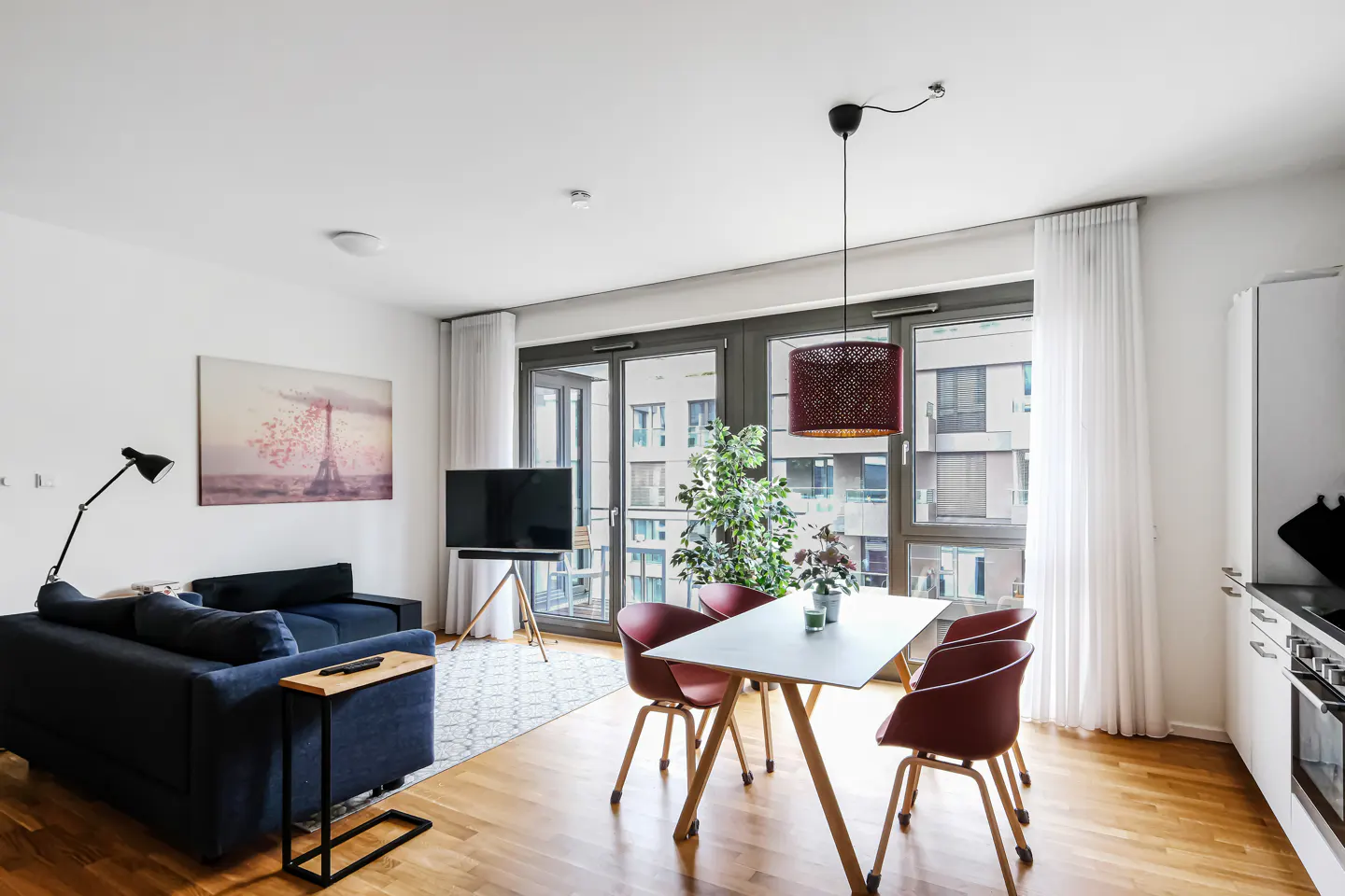 Bright apartment with a blue sofa, dining table with red chairs, and a TV. Large windows overlook a city view.