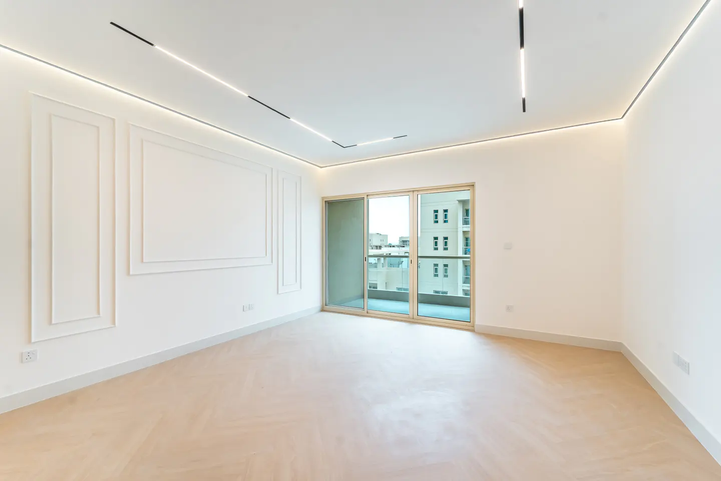 Bright, empty room with white walls, herringbone wood floor, and sliding glass doors to a balcony. Modern lighting fixtures on the ceiling.