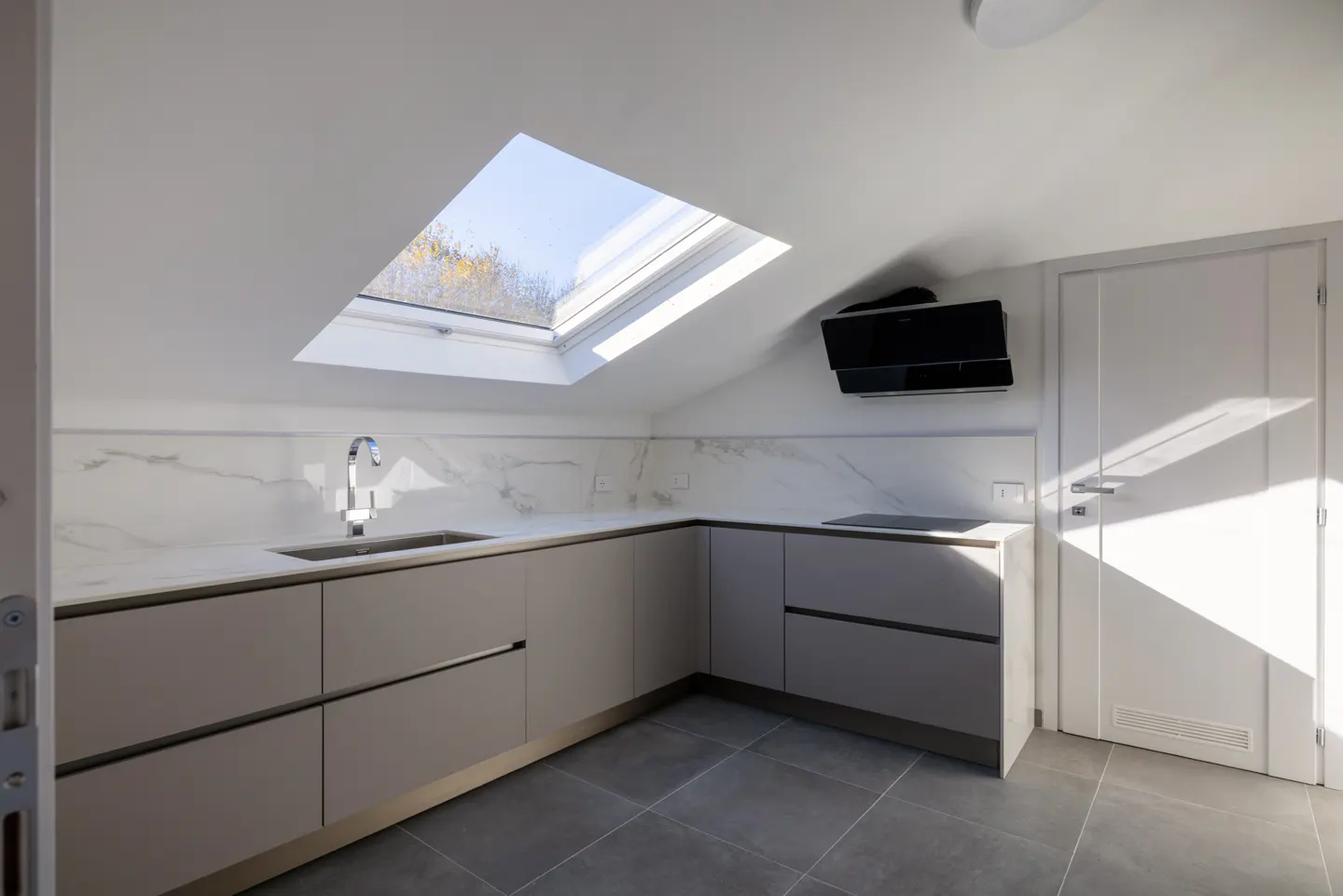 Bright, modern kitchen with gray cabinets, marble backsplash, and skylight. Black range hood and gray tile floor. White door on the right.